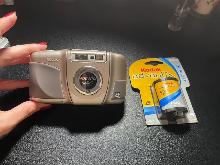 A hand holds a silver Kodak Advantix C200 film camera next to a packaged Kodak Advantix 200 film roll, both on a black surface under bright light.