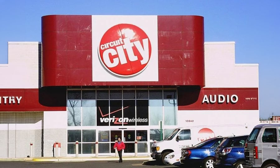 A person walks toward the entrance of a Circuit City store with a large red sign above the doors. Several cars are parked in front on a sunny day.
