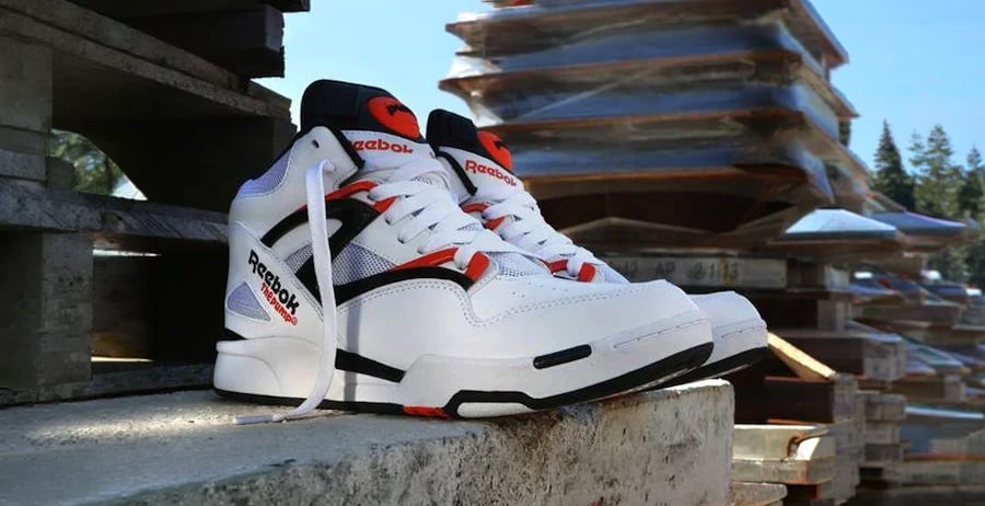 A pair of white Reebok sneakers with black and orange accents sits on a concrete slab outdoors, with stacked wooden pallets and trees visible in the background.