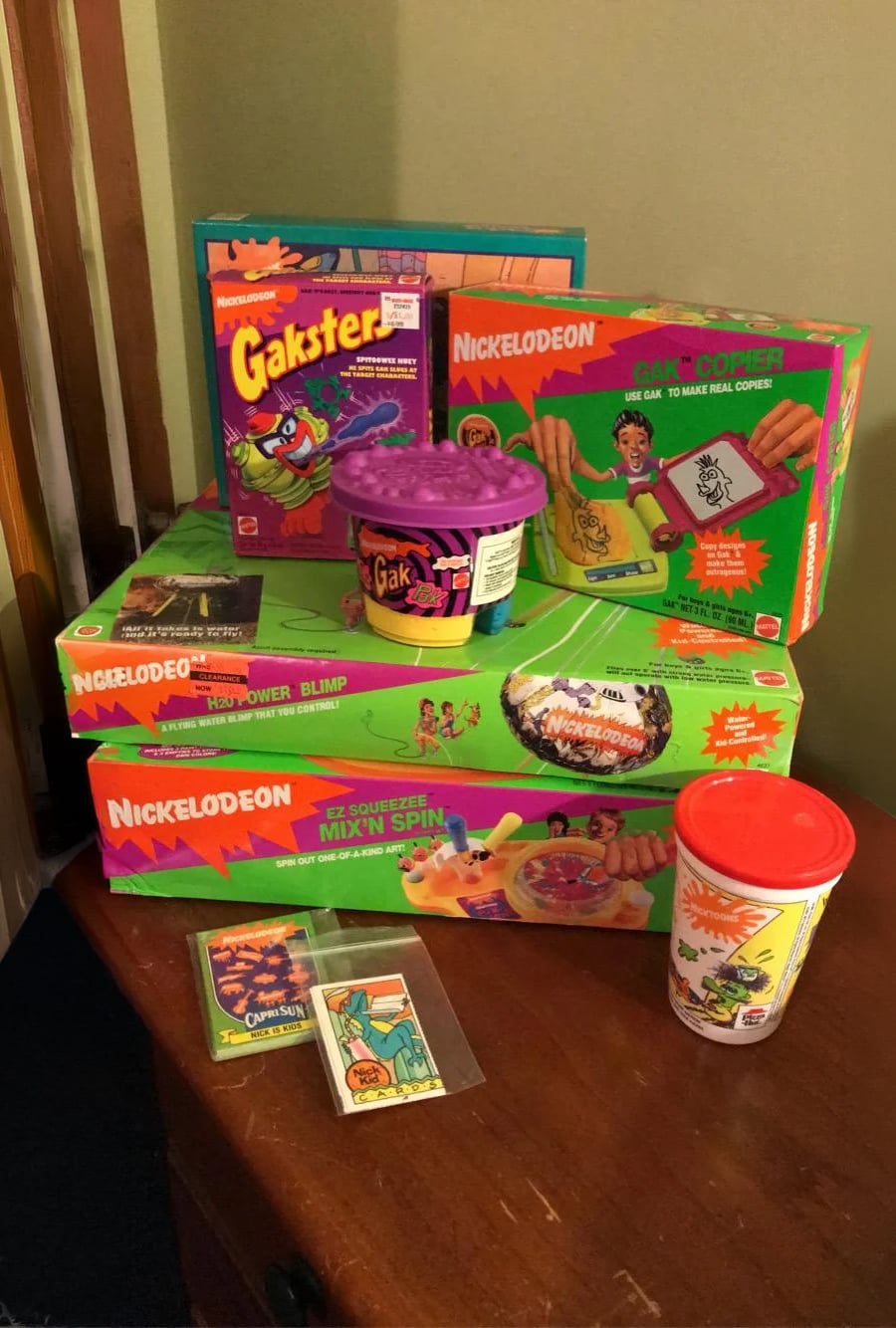 Several colorful, boxed Nickelodeon toys and kits, including Gak, Creepy Crawlers, and a cup, are stacked on a table against a beige wall. Small trading cards are placed at the front of the display.
