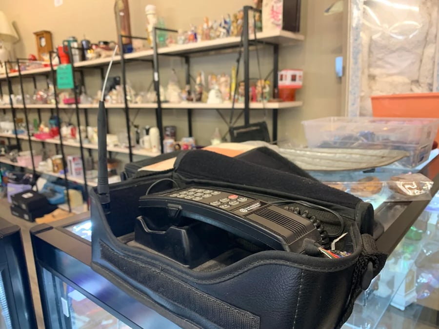 A vintage car phone with a long antenna sits in a black carrying case on a glass counter in a thrift store, with shelves of assorted figurines and items in the background.