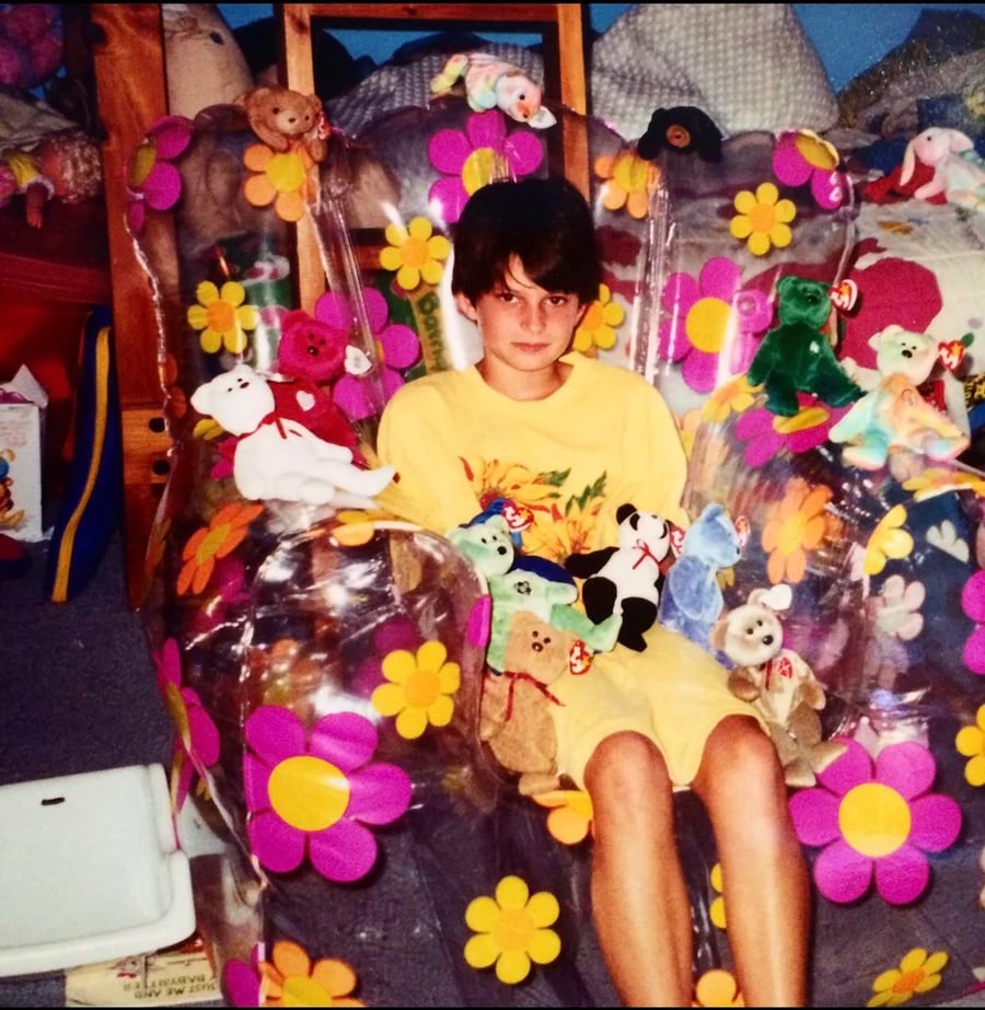 A child sits on a clear, flower-patterned inflatable chair surrounded by Beanie Babies in a cluttered room, wearing a yellow T-shirt and shorts, looking at the camera.