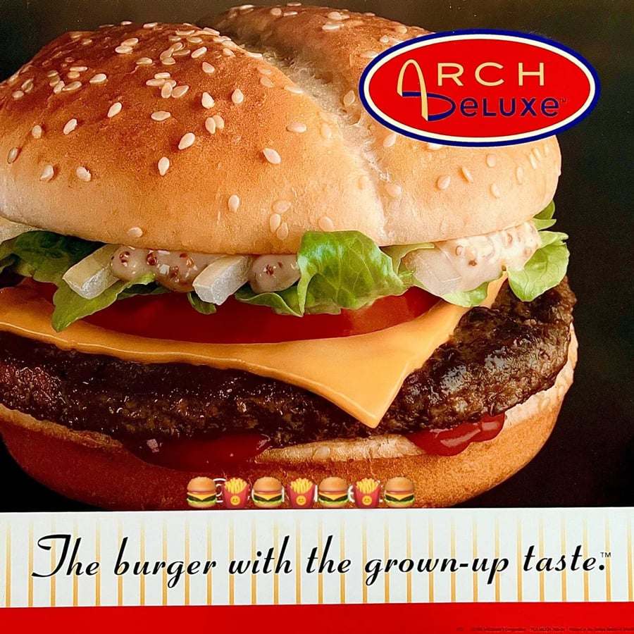 Close-up of a cheeseburger with lettuce, tomato, cheese, and sauce. The words “Arch Deluxe” and “The burger with the grown-up taste” are displayed, with burger and fries emojis underneath.