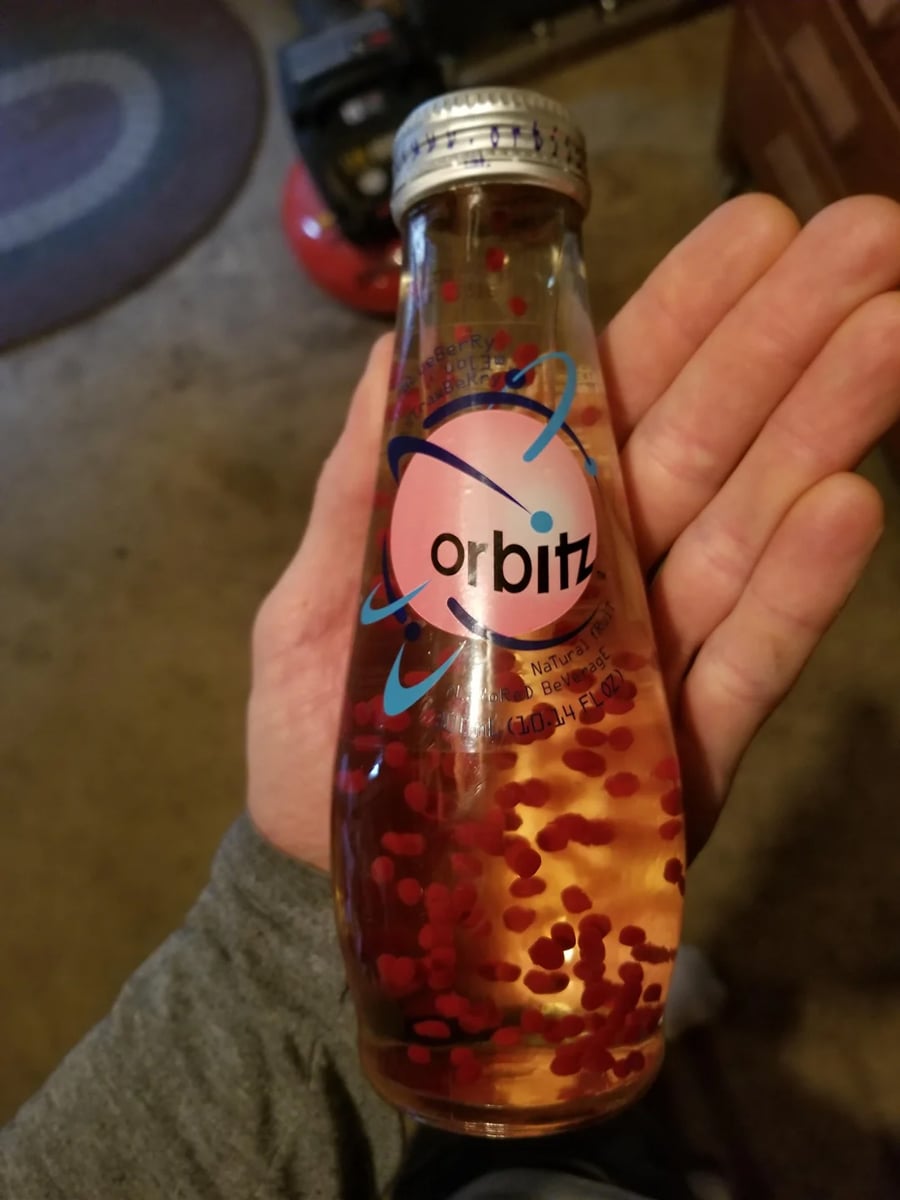 A hand holds a vintage glass bottle of Orbitz drink, with floating red gelatin balls suspended in a yellow liquid. The bottle has a silver cap and colorful label with the Orbitz logo.