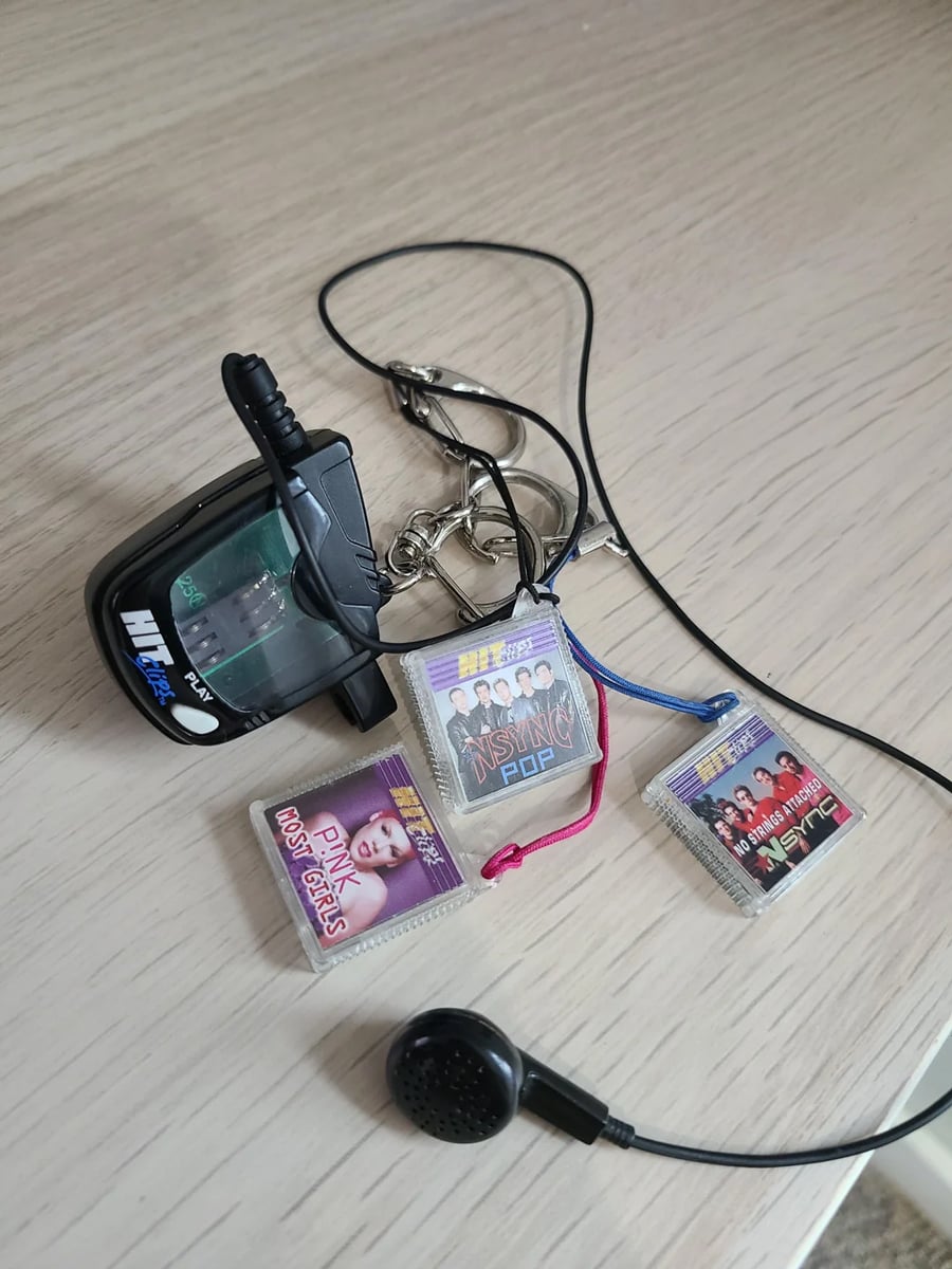 A small black music player with earphones, keys, and three tiny toy album covers for Pink, NSYNC, and the Backstreet Boys rest on a light wooden surface.