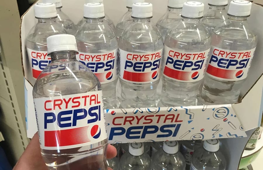A hand holds a bottle of Crystal Pepsi in front of a store display filled with more Crystal Pepsi bottles, all featuring clear soda and retro-style labels.