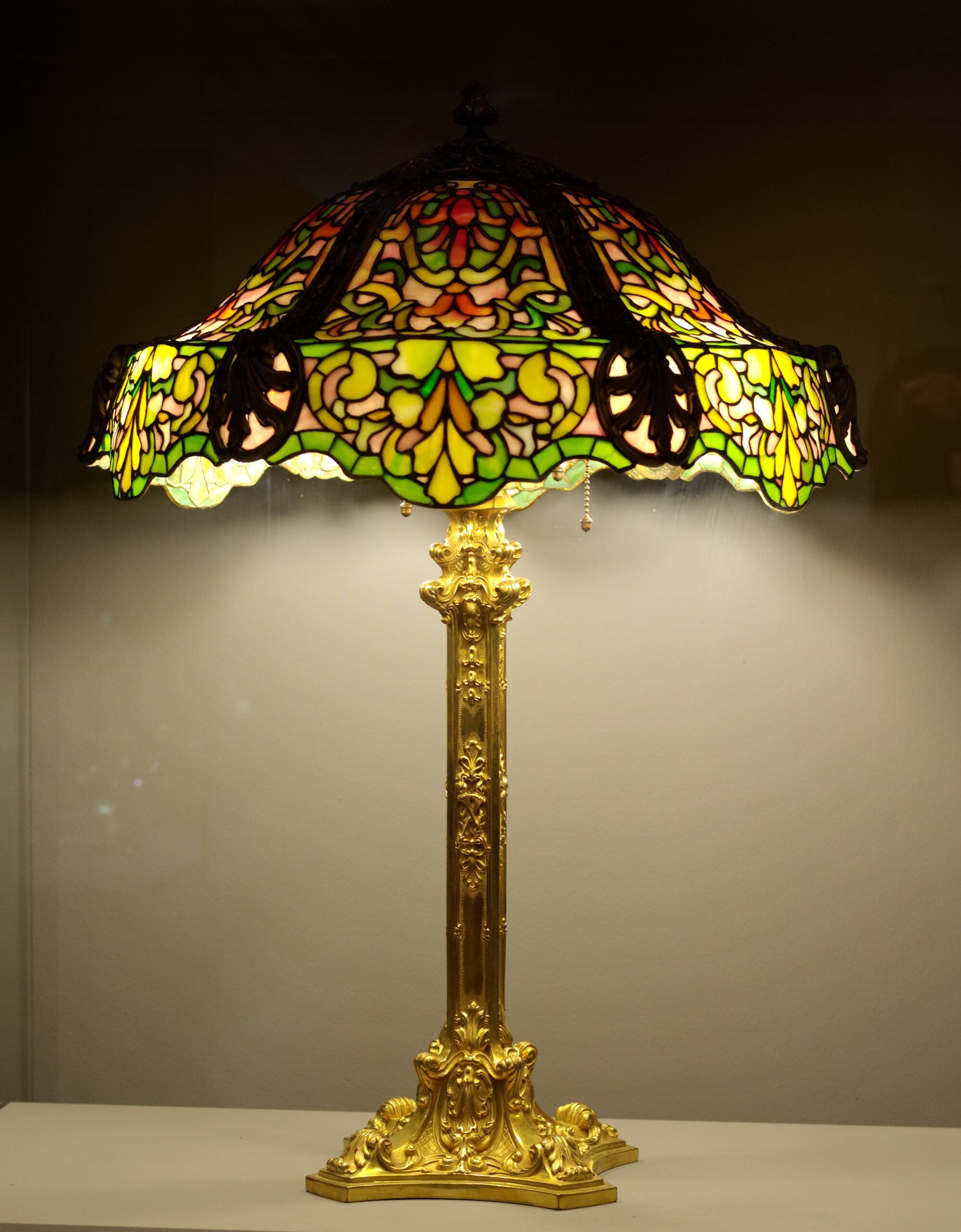 A Tiffany-style lamp with a colorful stained glass shade in green, yellow, and red patterns, and a detailed gold base, is turned on and displayed against a plain background.