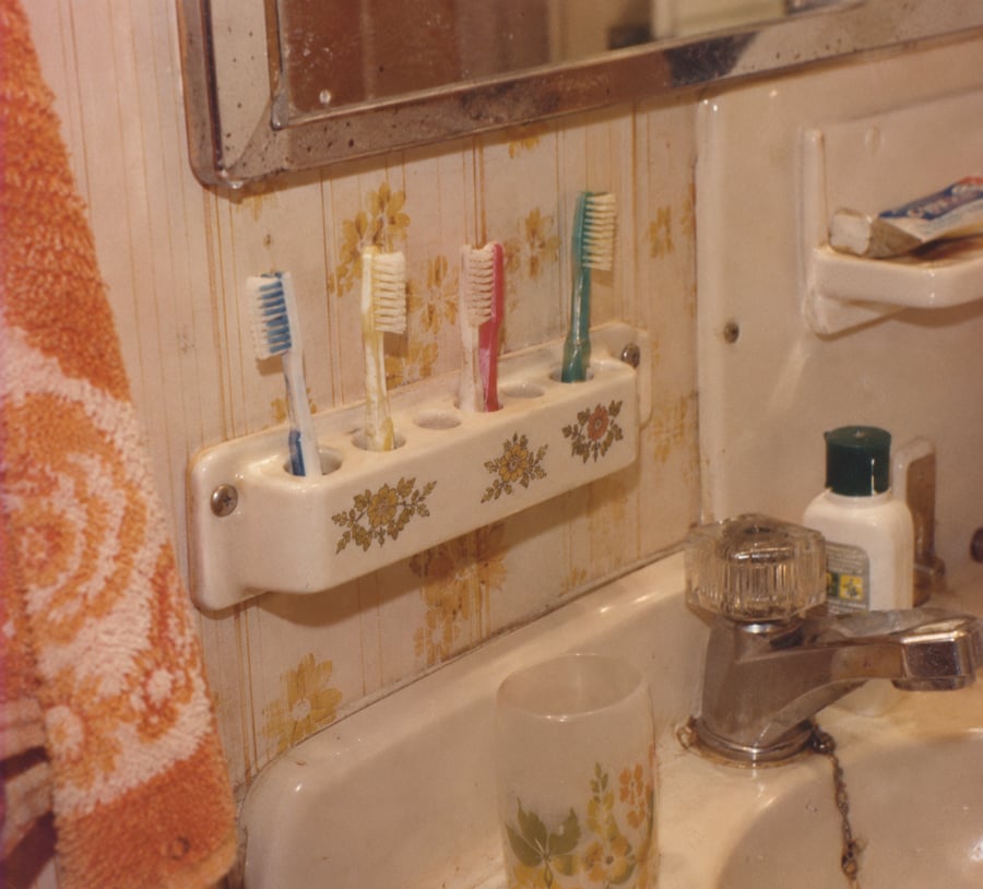 Four toothbrushes in a ceramic floral holder on a yellow floral wallpapered bathroom wall, next to a sink with a cup and toiletries, and a towel hanging nearby.
