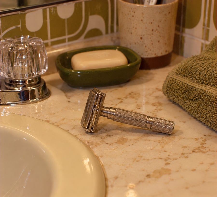 A metal safety razor lies on a marble bathroom counter near a sink, next to a bar of soap in a green dish, a glass faucet handle, a green towel, and a speckled cup holding toothbrushes.