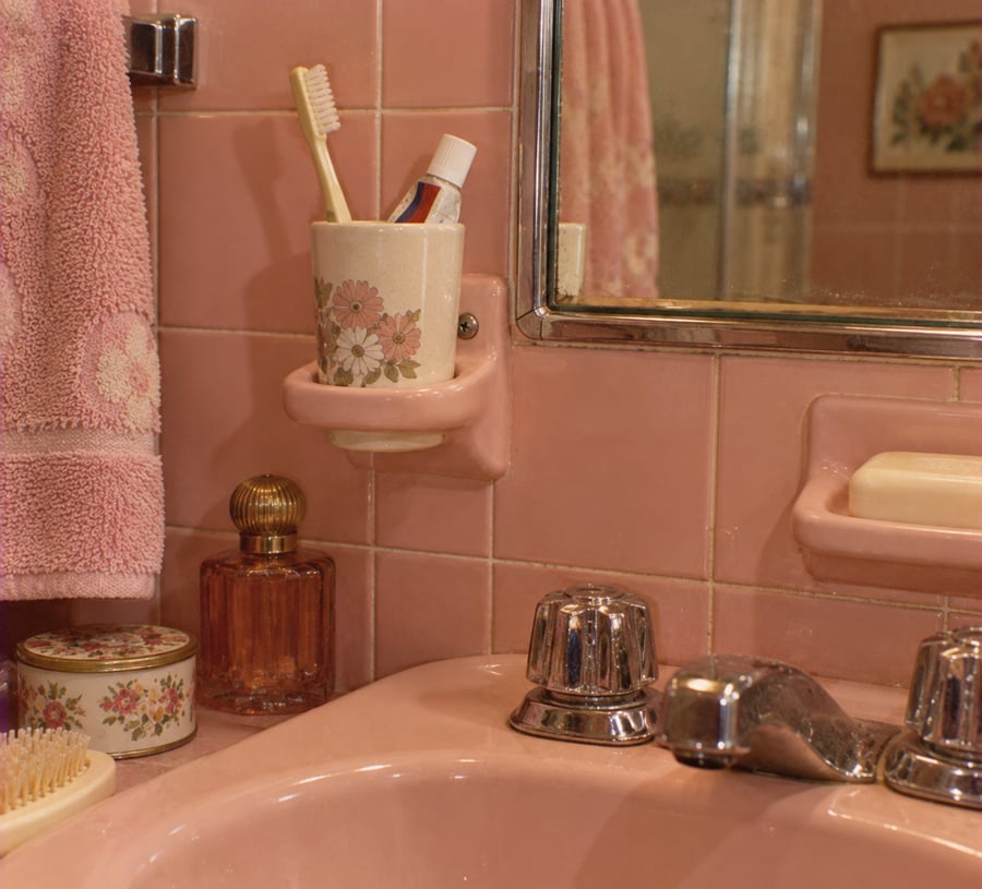 A vintage pink bathroom with a pink sink, floral toothbrush holder, toothpaste, floral towel, soap dish, perfume bottle, brush, and a wall mirror, all accented with floral patterns.