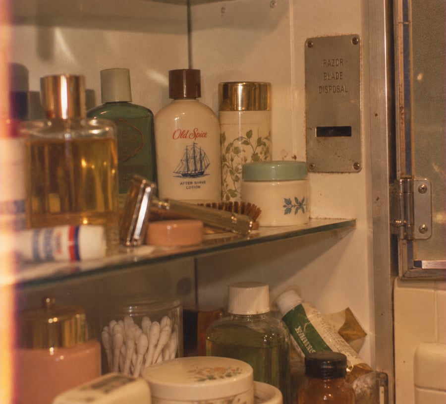 A close-up of a bathroom medicine cabinet with various vintage toiletries, including Old Spice after shave, jars, a razor, cotton swabs, toothpaste, and a metal razor blade disposal slot on the right.