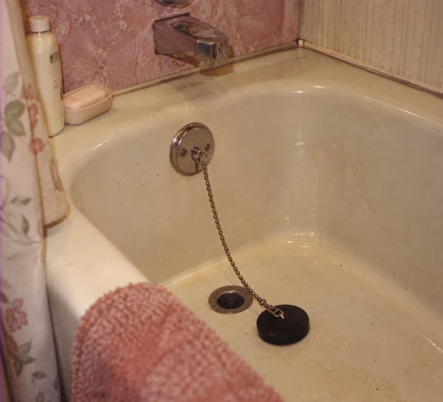 A worn bathtub with a pink towel, floral shower curtain, soap, a shampoo bottle, and a drain plug attached to a chain. The tub and tiles show visible signs of age and staining.