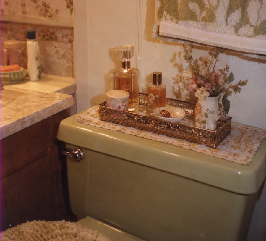 A vintage bathroom scene shows a green toilet with a decorative tray holding perfume bottles, a small vase of flowers, and a dish. A towel hangs above and toiletries sit on a marble counter nearby.