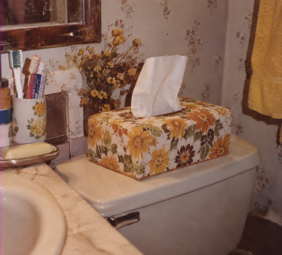 A floral tissue box sits on a toilet tank in a bathroom, next to dried flowers, toothbrushes in a cup, a yellow towel, and a bar of soap on a marble counter. The wallpaper features a small floral pattern.