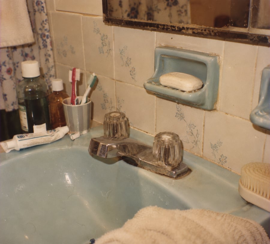 A vintage bathroom sink with two clear faucet handles, a bar of soap on a blue holder, toothbrushes in a cup, toothpaste, bottles, a brush, and a towel. The wall tiles have a small blue floral pattern.