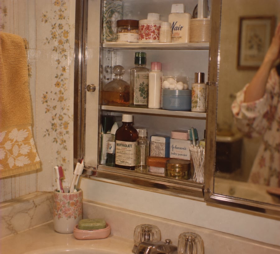 A bathroom sink with floral cups holding toothbrushes, bars of soap, and an open mirrored medicine cabinet displaying various bottles, jars, and personal care items. A person’s arm is partially visible in the reflection.