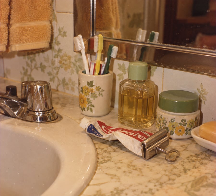 A bathroom sink with a floral cup holding toothbrushes, a toothpaste tube, two jars, a bar of soap, a faucet, and a hand towel hanging above, all on a marble countertop with floral tiles in the background.