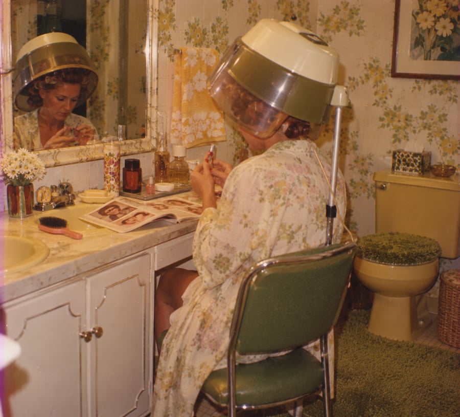 A woman in a floral robe sits at a vanity with a hair dryer hood on, reading a magazine in a vintage bathroom decorated with yellow and green floral wallpaper and matching green toilet cover and rug.