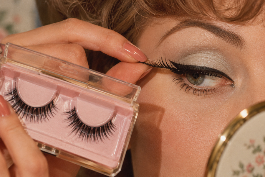 A person applies a false eyelash while holding a box of lashes, with one eye already made up and a small mirror visible in the foreground.
