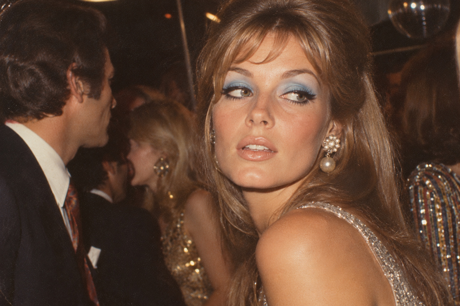 A woman with long hair, blue eyeshadow, pearl earrings, and a sparkly dress looks over her shoulder at a crowded, glamorous party.