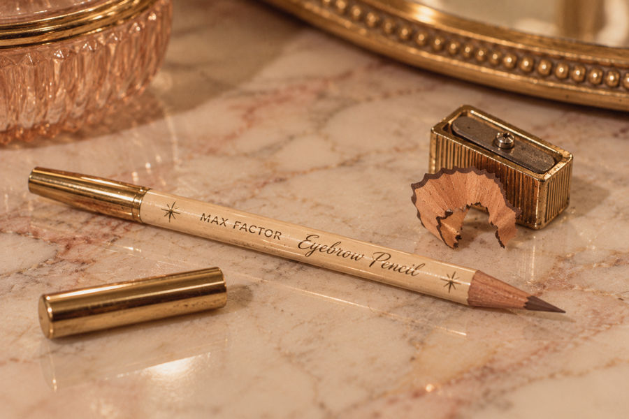 A Max Factor eyebrow pencil, sharpener, and cap rest on a marble surface with a gold mirror and a small shaved pencil curl nearby.