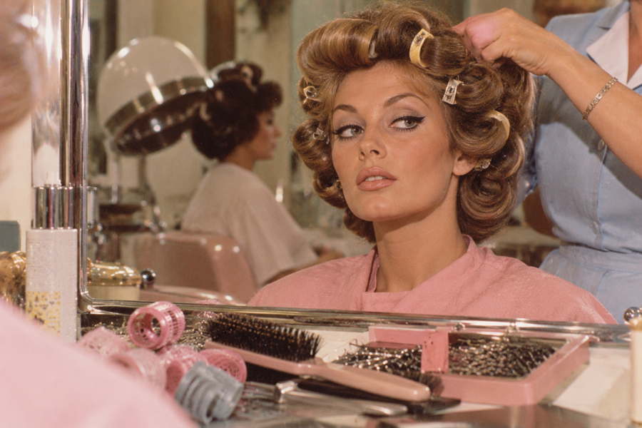 A woman in a pink robe sits at a salon station with hair curlers while a stylist adjusts her hair. Salon tools and brushes are visible on the counter. Another woman is blurred in the background.