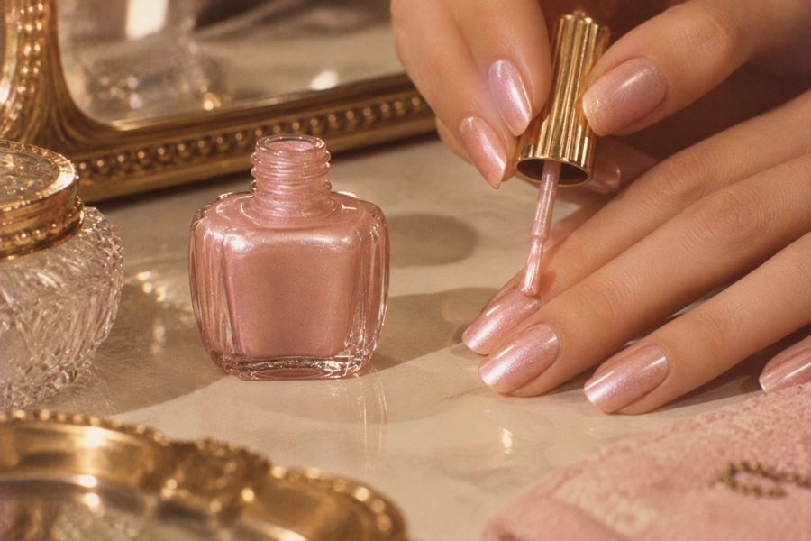 A person applies shimmery pink nail polish to their fingernails. An open nail polish bottle, vintage mirror, and glass container are on a marble surface nearby.