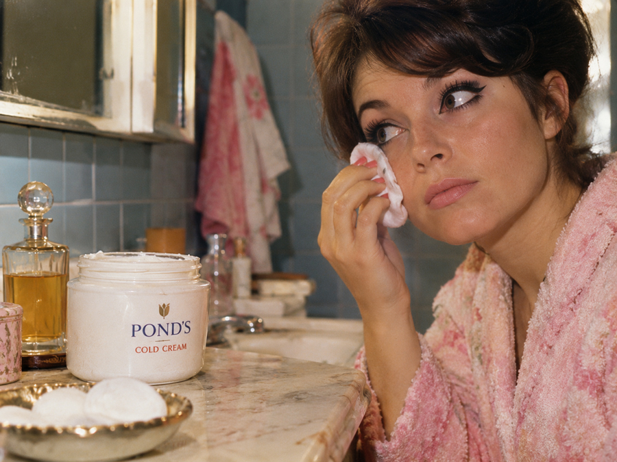 A woman in a pink robe applies Pond’s cold cream to her face with a cotton pad in a bathroom with blue tiles and vintage decor.