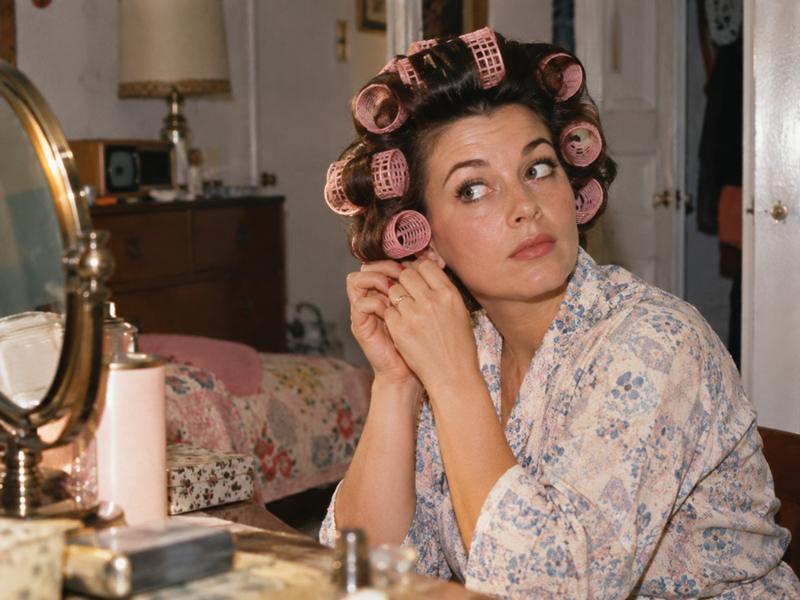 Woman wearing pink hair rollers and a floral robe sits at a vanity, adjusting an earring and looking to the side. The room contains a bed, dresser, lamp, and mirror.