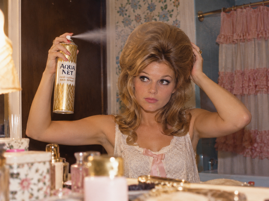 A woman with voluminous hair sprays Aqua Net hairspray while looking in a bathroom mirror, surrounded by vintage decor and beauty products.