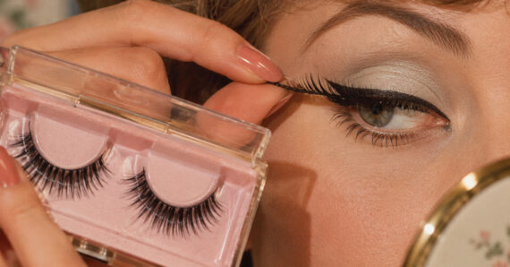A person applies a false eyelash to their upper eyelid while holding a pink case containing another pair of false eyelashes.