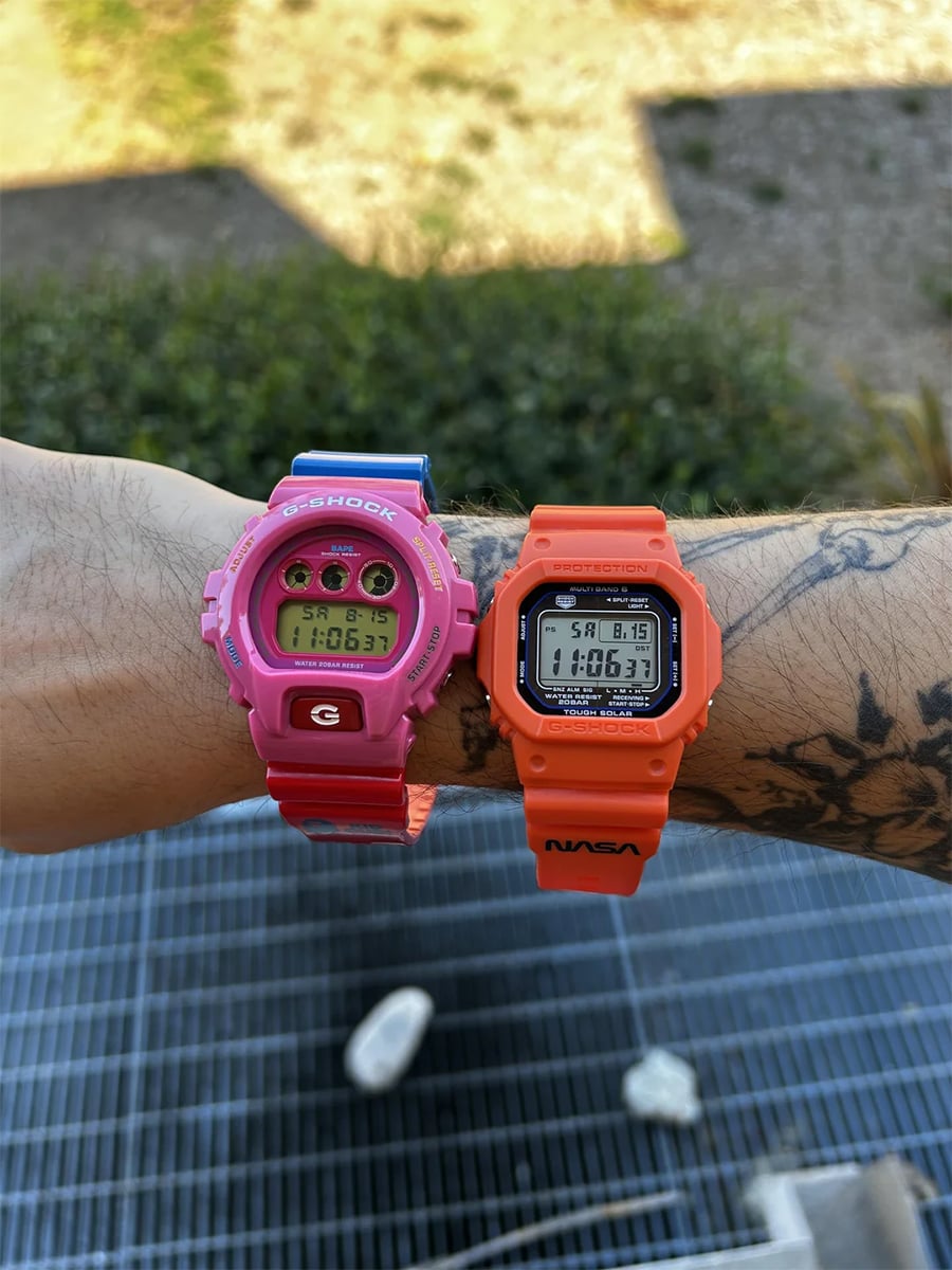 Two tattooed wrists wearing digital watches: a pink G-Shock on the left wrist and an orange NASA G-Shock on the right, both displaying the time 10:06. Outdoor background with greenery and gravel.