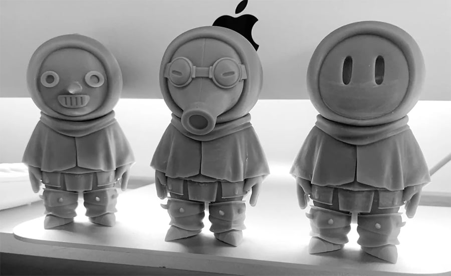 Three stylized astronaut figurines with distinct faces—one smiling, one with a gas mask, and one with a toothy grin—stand in front of a lit computer monitor with an Apple logo.