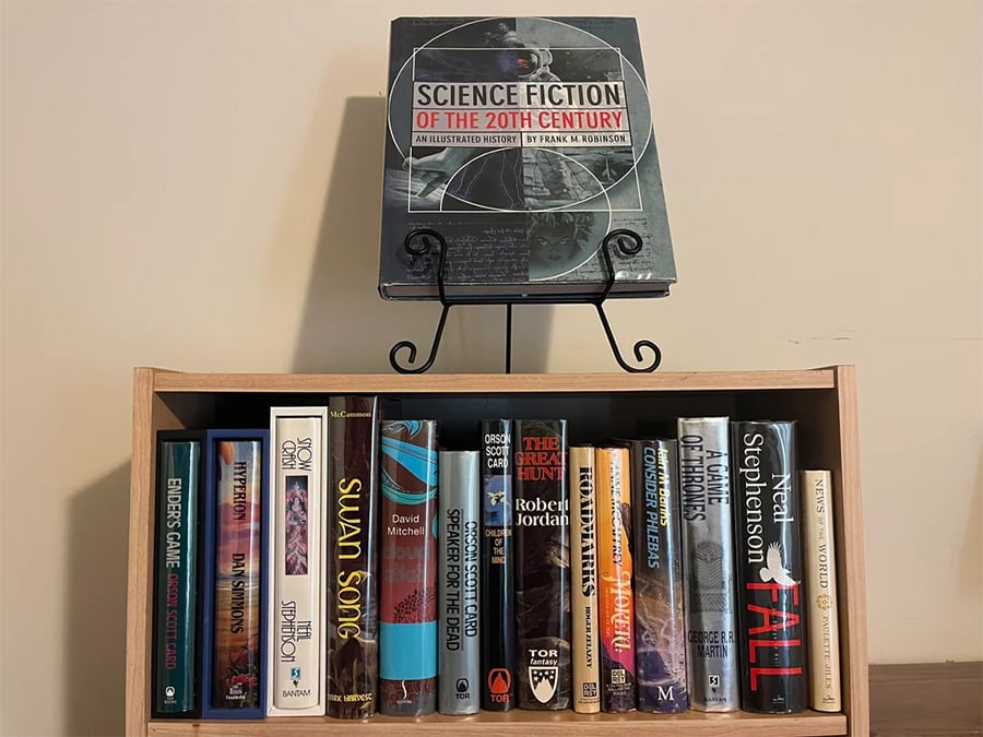 A bookshelf displays science fiction and fantasy novels, with a large book titled “Science Fiction of the 20th Century” on a stand above the shelf.