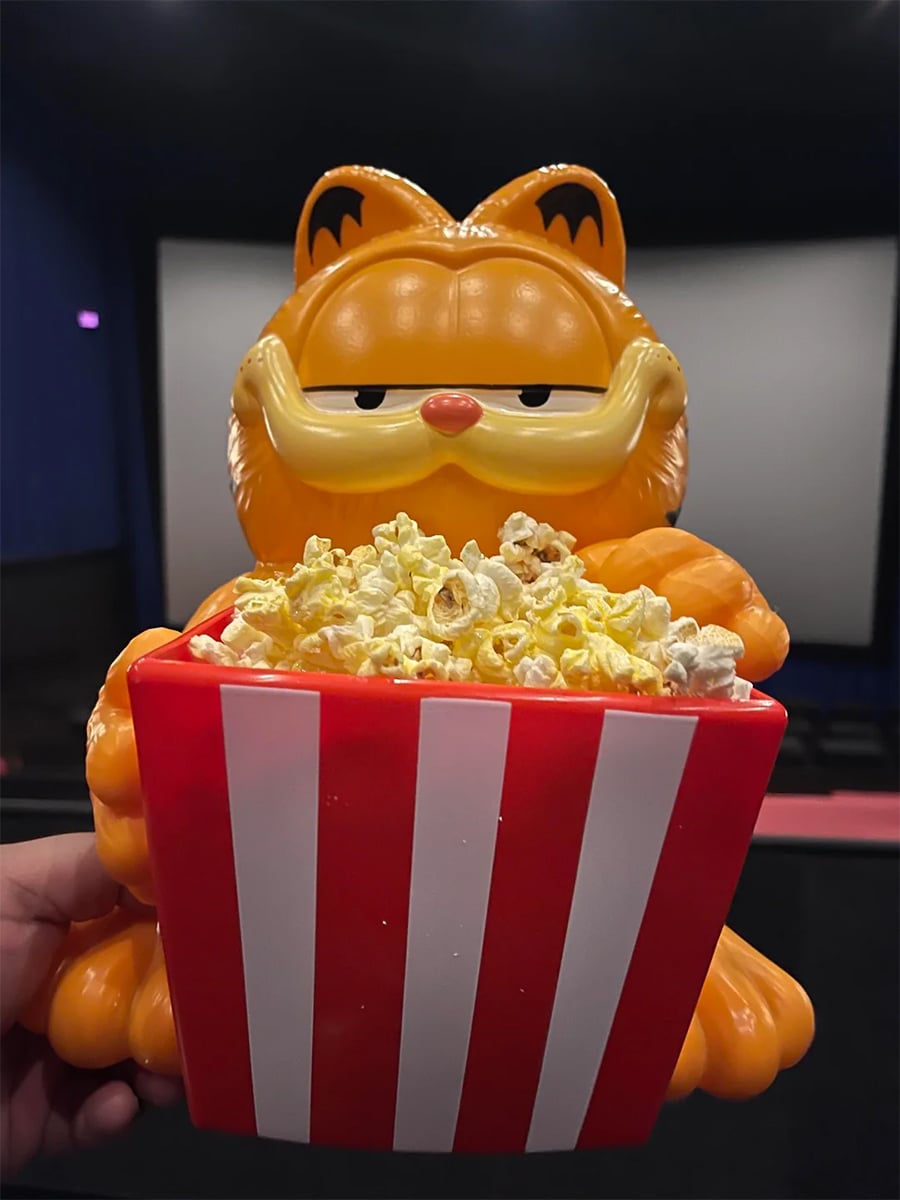 A Garfield popcorn bucket filled with popcorn is held up in a movie theater, with a blank movie screen visible in the background.