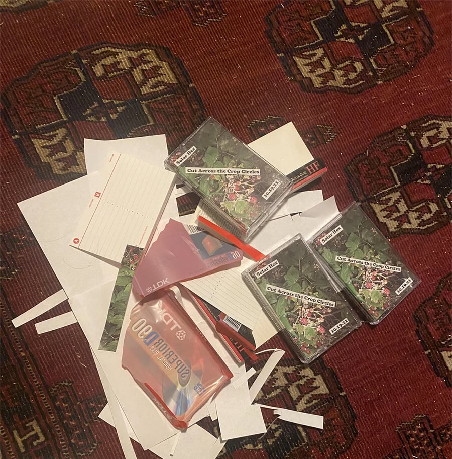 A pile of white paper scraps, cut-up cards, and four cassette tapes titled “Cut Across the Cry Circle” are scattered on a patterned red rug.