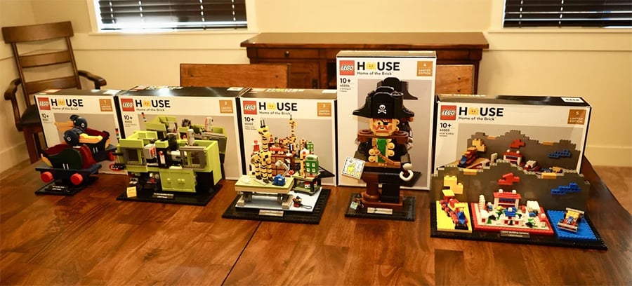 Five LEGO sets from the House series, each built and displayed in front of its box, are arranged on a wooden table with chairs and a sideboard in the background.