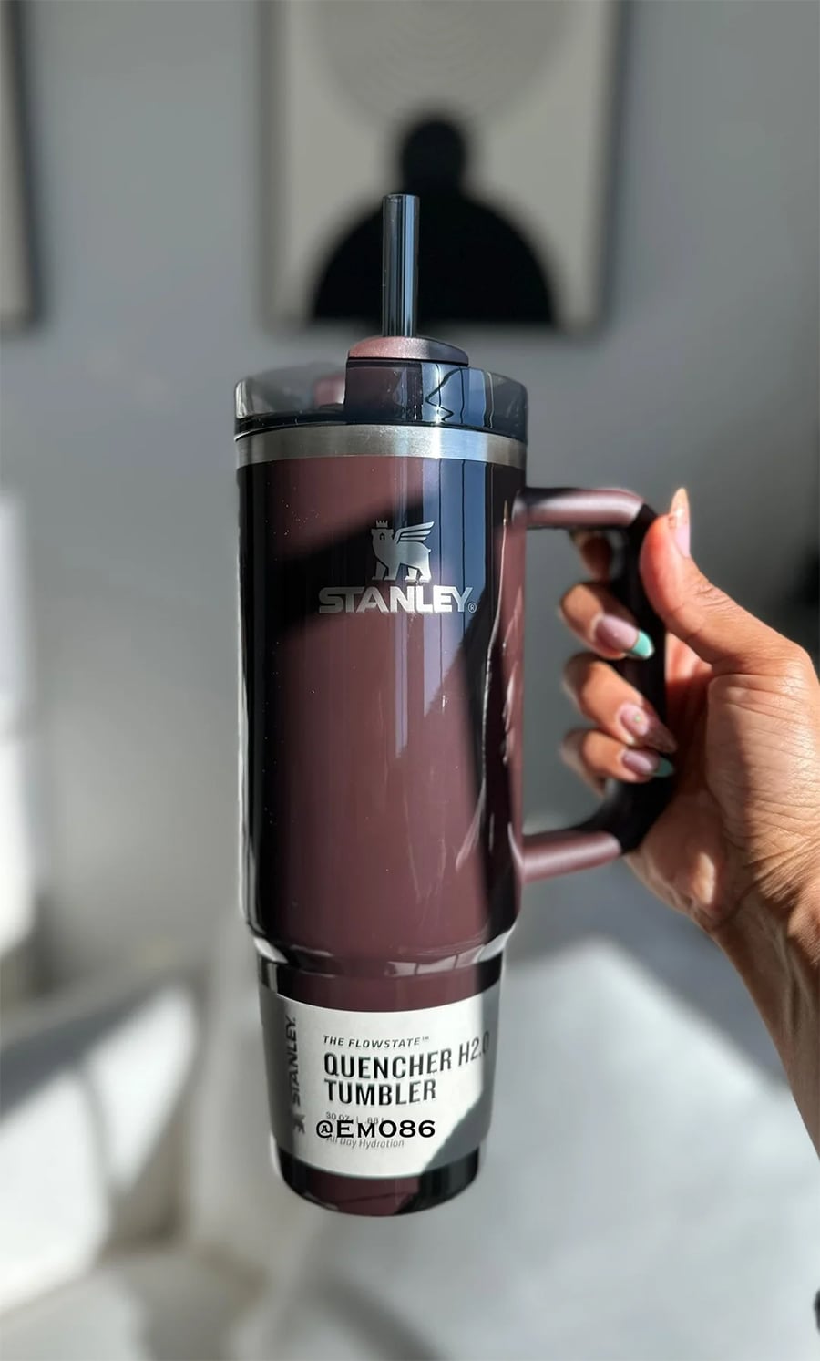 A hand holding a maroon Stanley Quencher H2.0 Tumbler with a straw. The tumbler is shiny and well-lit, and the background is softly blurred with neutral tones.