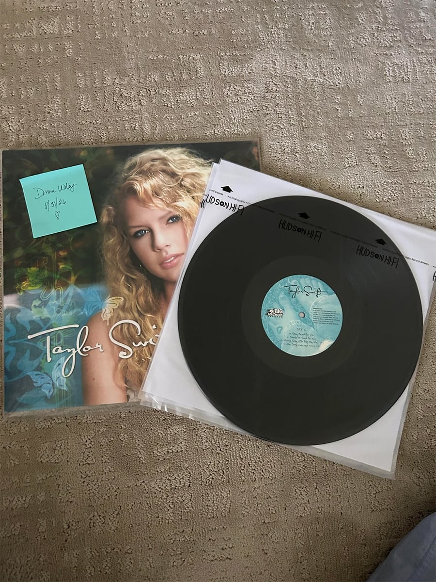 A vinyl record and its cover featuring Taylor Swift’s self-titled album are on a carpet. A blue sticky note on the cover reads, “Dear Walker, 11/21/21 Hi!” with a heart.