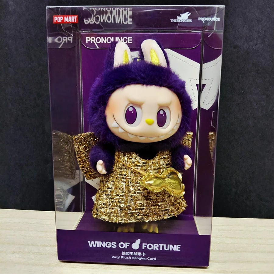 A vinyl plush figurine in a gold robe with a purple fuzzy hood and animal ears, packaged in a clear display box labeled “WINGS OF FORTUNE” by Pop Mart and Pronounce.