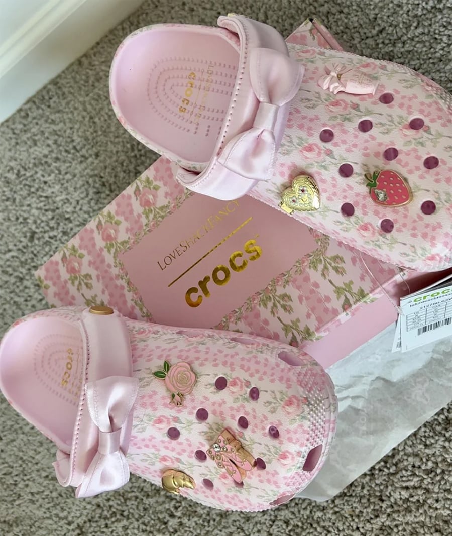 Pink Crocs with bows, heart and strawberry charms, resting on a decorative pink floral Crocs box atop a beige carpet.