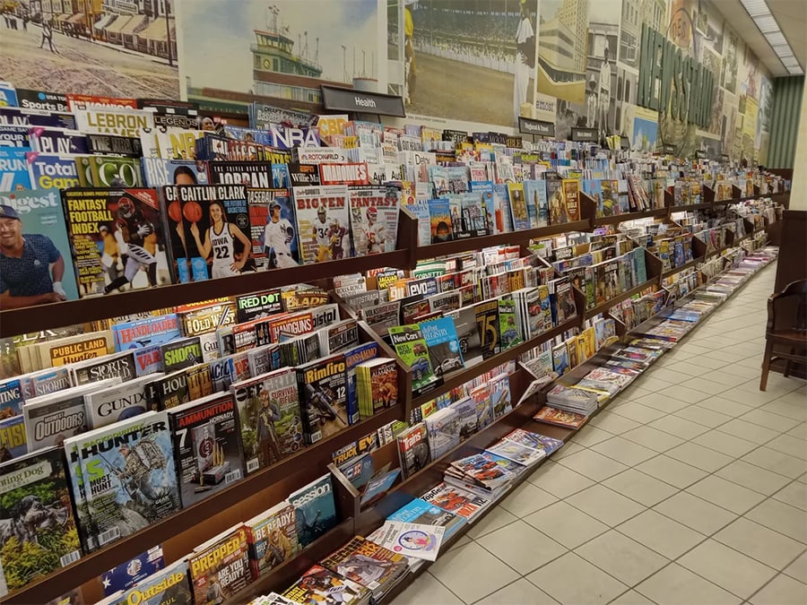 A bookstore magazine section displays rows of colorful magazines on wooden shelves, featuring topics like sports, news, entertainment, fashion, and hobbies. The tiled floor and wall murals are visible in the background.