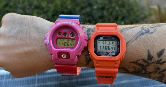 A tattooed arm wears two digital G-Shock watches: one pink and round, the other orange and square, both displaying the same time and date. Green plants are blurred in the background.