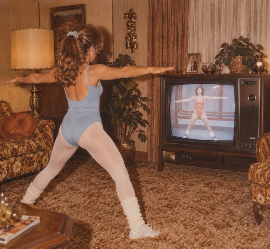 A woman in workout clothes does aerobics in a living room while following a fitness instructor on a vintage television.