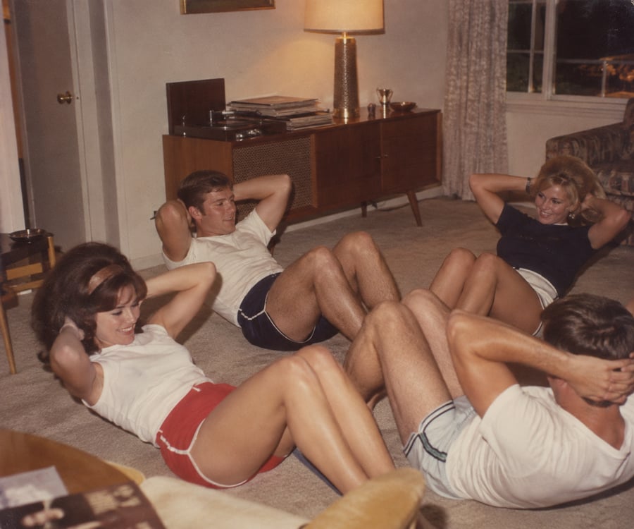 Four adults, two men and two women, wearing shorts and t-shirts, do sit-ups together on a carpeted floor in a living room with a lamp, cabinet, and window in the background.