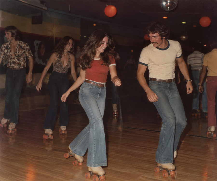 People roller skating on a wooden rink, wearing 1970s-style clothes. A woman in a red top and a man in a white shirt skate together, smiling. Other skaters are visible in the background.
