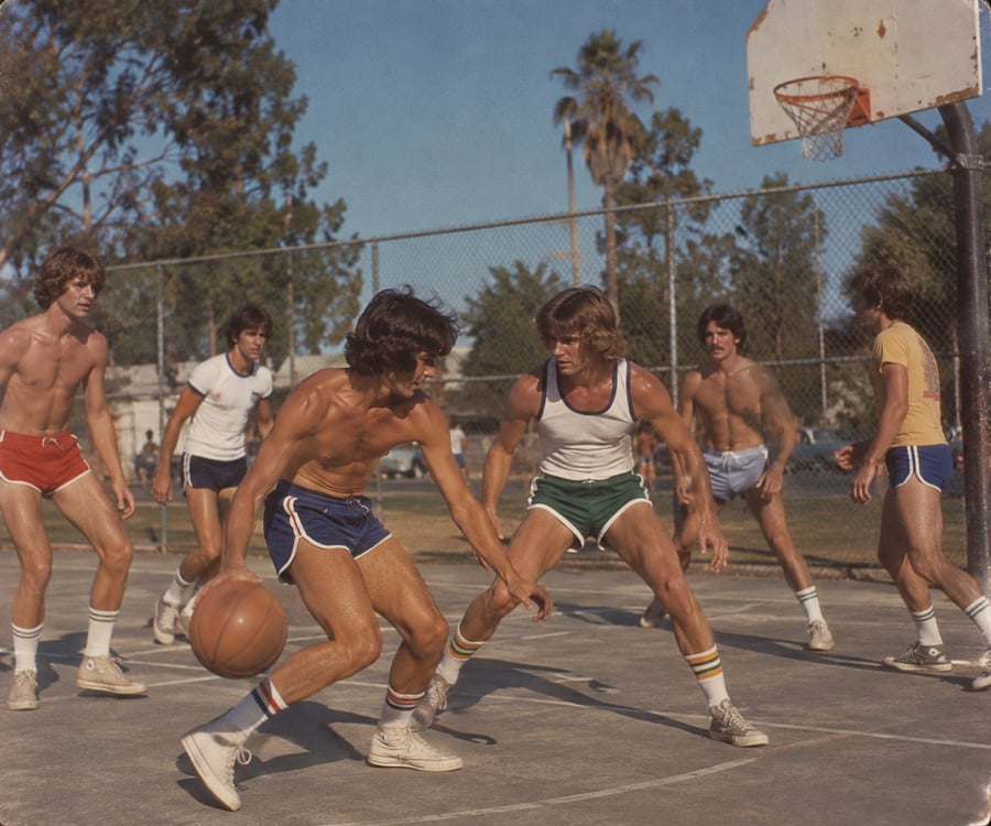 Six young men in 1970s athletic wear play basketball outdoors on a sunny day. One dribbles the ball while others guard or watch. Trees and a chain-link fence are visible in the background.