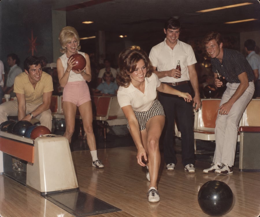 A woman bowls while four friends, two women and two men, watch and smile in a retro bowling alley. The group appears to be having fun together.