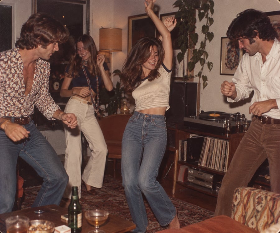 Four people dance energetically in a cozy, warmly lit living room with a record player, drinks on the table, plants, and vintage decor.