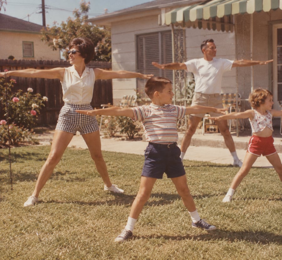A family of four does exercises together in a backyard, arms outstretched, on a sunny day. Two adults and two children wear summer clothes and stand on grass near a house.